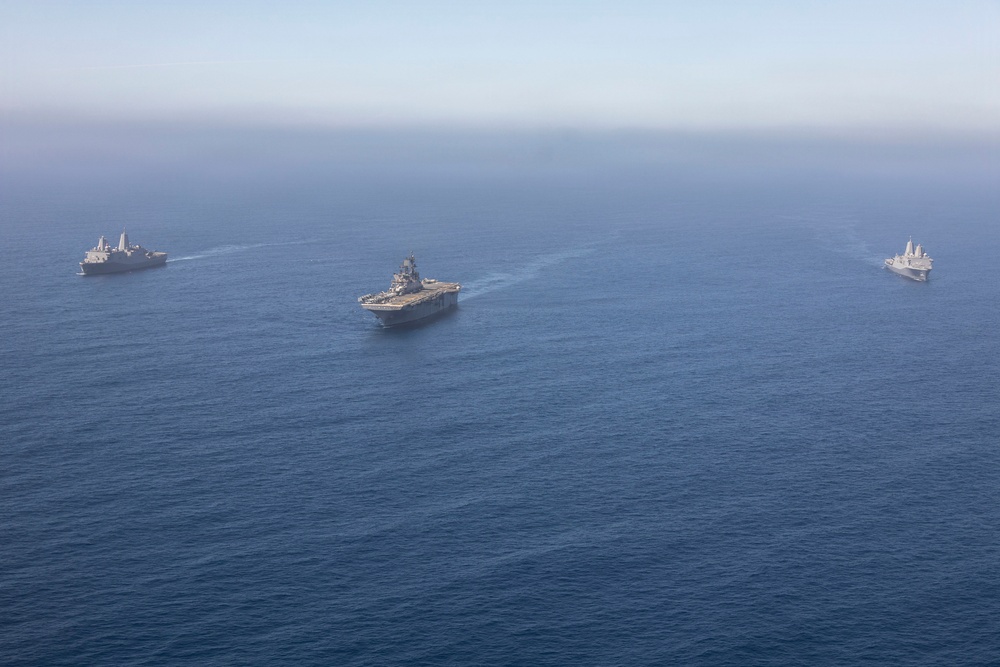 An aerial photograph shows three large naval vessels sailing in formation across open blue ocean waters. The central ship appears to be an amphibious assault ship or aircraft carrier, flanked by two smaller amphibious transport dock vessels on either side. The ships are leaving visible wake trails as they travel in a coordinated formation.