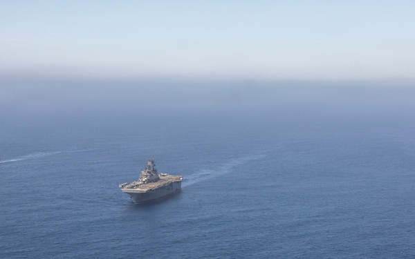 An aerial photograph shows three large naval vessels sailing in formation across open blue ocean waters. The central ship appears to be an amphibious assault ship or aircraft carrier, flanked by two smaller amphibious transport dock vessels on either side. The ships are leaving visible wake trails as they travel in a coordinated formation.