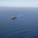 An aerial photograph shows three large naval vessels sailing in formation across open blue ocean waters. The central ship appears to be an amphibious assault ship or aircraft carrier, flanked by two smaller amphibious transport dock vessels on either side. The ships are leaving visible wake trails as they travel in a coordinated formation.