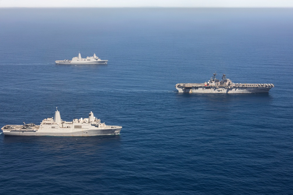 An aerial photograph shows three naval vessels sailing in formation across open blue ocean waters. The formation includes a large amphibious assault ship or aircraft carrier on the right, a San Antonio-class amphibious transport dock ship in the foreground left, and a smaller warship in the background. The ships appear to be conducting a joint naval exercise or transit operation.