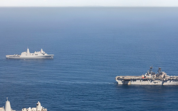 An aerial photograph shows three naval vessels sailing in formation across open blue ocean waters. The formation includes a large amphibious assault ship or aircraft carrier on the right, a San Antonio-class amphibious transport dock ship in the foreground left, and a smaller warship in the background. The ships appear to be conducting a joint naval exercise or transit operation.