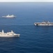 An aerial photograph shows three naval vessels sailing in formation across open blue ocean waters. The formation includes a large amphibious assault ship or aircraft carrier on the right, a San Antonio-class amphibious transport dock ship in the foreground left, and a smaller warship in the background. The ships appear to be conducting a joint naval exercise or transit operation.