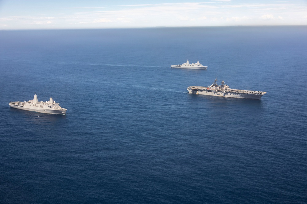 An aerial photograph shows three U.S. Navy warships sailing in formation across open blue ocean. The formation includes one large amphibious assault ship (resembling a Wasp-class or America-class LHA/LHD) with a flat flight deck visible in the center-right, flanked by two San Antonio-class amphibious transport dock (LPD) ships. The ships appear to be conducting a routine underway transit or naval exercise.