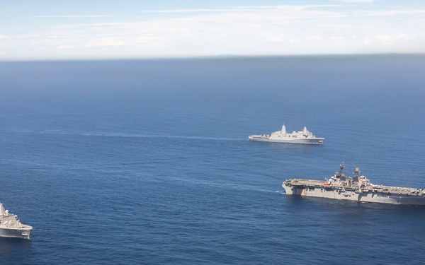 An aerial photograph shows three U.S. Navy warships sailing in formation across open blue ocean. The formation includes one large amphibious assault ship (resembling a Wasp-class or America-class LHA/LHD) with a flat flight deck visible in the center-right, flanked by two San Antonio-class amphibious transport dock (LPD) ships. The ships appear to be conducting a routine underway transit or naval exercise.
