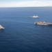 An aerial photograph shows three U.S. Navy warships sailing in formation across open blue ocean. The formation includes one large amphibious assault ship (resembling a Wasp-class or America-class LHA/LHD) with a flat flight deck visible in the center-right, flanked by two San Antonio-class amphibious transport dock (LPD) ships. The ships appear to be conducting a routine underway transit or naval exercise.