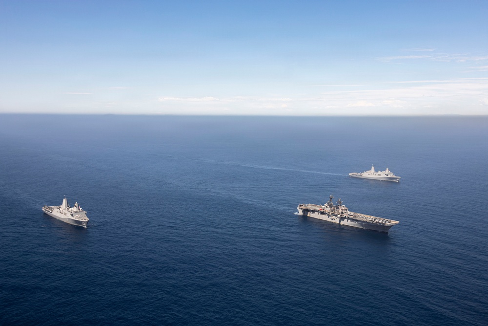 An aerial photograph shows three naval vessels sailing in formation across open blue ocean under a partly cloudy sky. The central and largest vessel appears to be an amphibious assault ship with a flat flight deck, flanked by two smaller amphibious transport dock or landing platform dock vessels. The ships are maintaining a triangular formation with significant spacing between them.