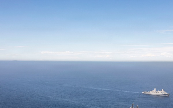 An aerial photograph shows three naval vessels sailing in formation across open blue ocean under a partly cloudy sky. The central and largest vessel appears to be an amphibious assault ship with a flat flight deck, flanked by two smaller amphibious transport dock or landing platform dock vessels. The ships are maintaining a triangular formation with significant spacing between them.