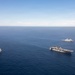 An aerial photograph shows three naval vessels sailing in formation across open blue ocean under a partly cloudy sky. The central and largest vessel appears to be an amphibious assault ship with a flat flight deck, flanked by two smaller amphibious transport dock or landing platform dock vessels. The ships are maintaining a triangular formation with significant spacing between them.