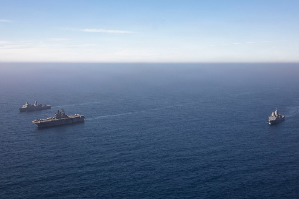 An aerial photograph shows three naval vessels sailing in formation across open blue ocean. The central and largest vessel appears to be an amphibious assault ship or aircraft carrier, flanked by two smaller warships. The ships are traveling in a loose triangular formation under clear skies.