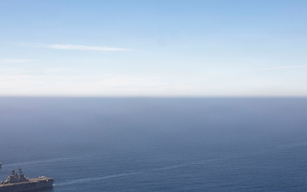 An aerial photograph shows three naval vessels sailing in formation across open blue ocean. The central and largest vessel appears to be an amphibious assault ship or aircraft carrier, flanked by two smaller warships. The ships are traveling in a loose triangular formation under clear skies.