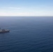 An aerial photograph shows three naval vessels sailing in formation across open blue ocean. The central and largest vessel appears to be an amphibious assault ship or aircraft carrier, flanked by two smaller warships. The ships are traveling in a loose triangular formation under clear skies.