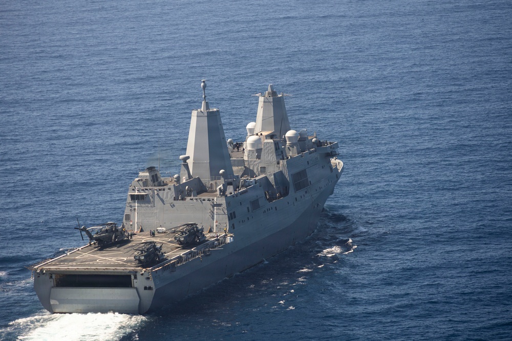 An aerial photograph of a large US Navy amphibious transport dock (LPD) ship underway in open blue ocean. The vessel features a flight deck at the stern with multiple helicopters and military vehicles visible. The ship displays characteristic stealth-angled superstructure consistent with the San Antonio-class LPD design.