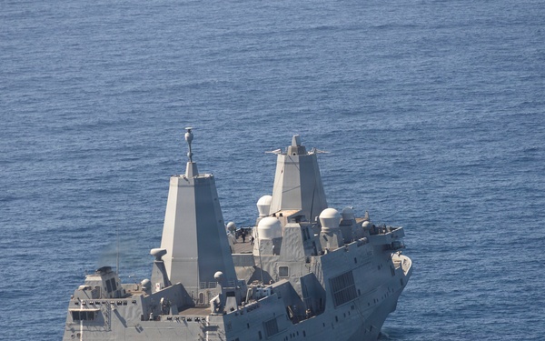 An aerial photograph of a large US Navy amphibious transport dock (LPD) ship underway in open blue ocean. The vessel features a flight deck at the stern with multiple helicopters and military vehicles visible. The ship displays characteristic stealth-angled superstructure consistent with the San Antonio-class LPD design.