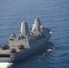 An aerial photograph of a large US Navy amphibious transport dock (LPD) ship underway in open blue ocean. The vessel features a flight deck at the stern with multiple helicopters and military vehicles visible. The ship displays characteristic stealth-angled superstructure consistent with the San Antonio-class LPD design.
