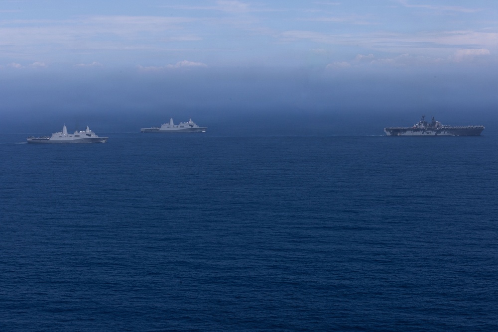 Three naval vessels are sailing in formation across open blue ocean under a hazy, overcast sky. The rightmost vessel appears to be a large amphibious assault ship or aircraft carrier, while the two vessels to the left appear to be amphibious transport dock (LPD-type) ships. The ships are spaced apart in a loose formation, suggesting a coordinated naval exercise or transit operation.