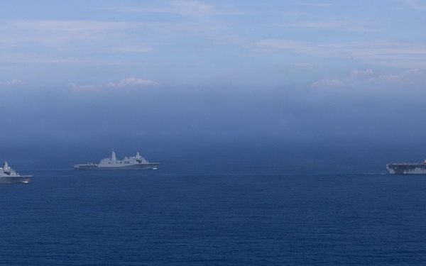 Three naval vessels are sailing in formation across open blue ocean under a hazy, overcast sky. The rightmost vessel appears to be a large amphibious assault ship or aircraft carrier, while the two vessels to the left appear to be amphibious transport dock (LPD-type) ships. The ships are spaced apart in a loose formation, suggesting a coordinated naval exercise or transit operation.