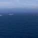 Three naval vessels are sailing in formation across open blue ocean under a hazy, overcast sky. The rightmost vessel appears to be a large amphibious assault ship or aircraft carrier, while the two vessels to the left appear to be amphibious transport dock (LPD-type) ships. The ships are spaced apart in a loose formation, suggesting a coordinated naval exercise or transit operation.