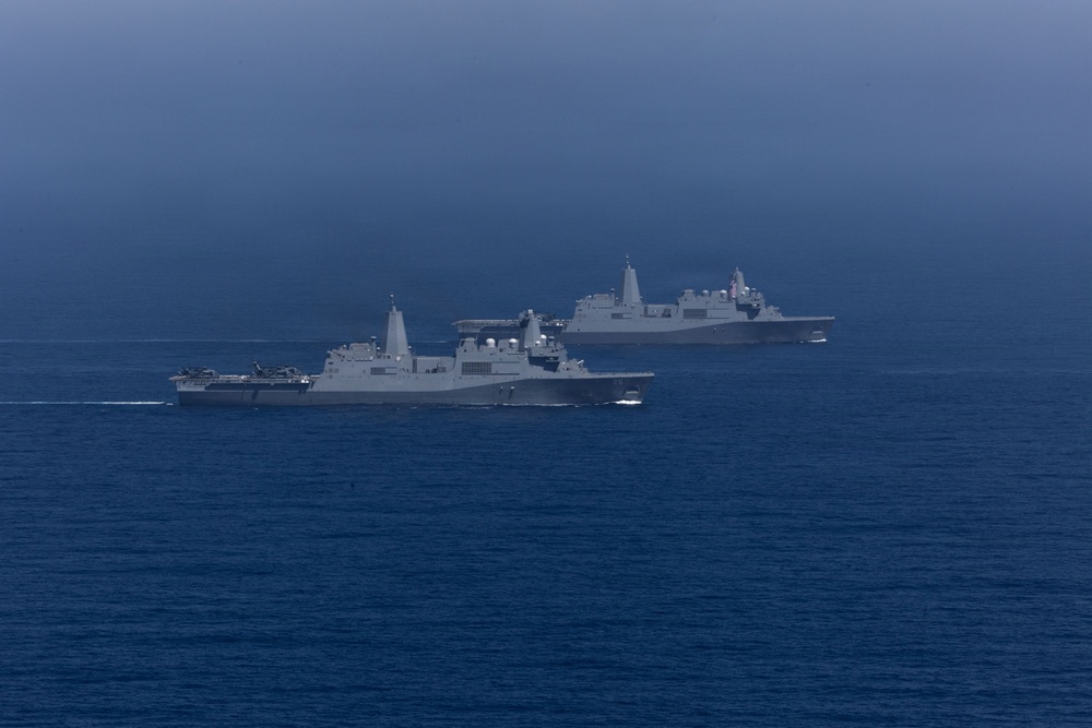 Two large gray naval warships are underway in open ocean under an overcast sky. Both vessels appear to be amphibious transport dock (LPD) class ships, sailing in a loose formation on calm, deep blue water. The image was taken from a distance, likely from a third vessel or aerial platform.