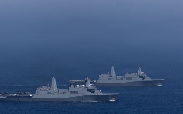 Two large gray naval warships are underway in open ocean under an overcast sky. Both vessels appear to be amphibious transport dock (LPD) class ships, sailing in a loose formation on calm, deep blue water. The image was taken from a distance, likely from a third vessel or aerial platform.
