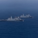 Two large gray naval warships are underway in open ocean under an overcast sky. Both vessels appear to be amphibious transport dock (LPD) class ships, sailing in a loose formation on calm, deep blue water. The image was taken from a distance, likely from a third vessel or aerial platform.
