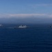 Three gray military naval vessels are sailing in formation across open deep-blue ocean waters. The ships appear to be modern warships, likely belonging to the same naval force or conducting a joint exercise. The scene is photographed from a distance, with a partially cloudy horizon and blue sky in the background.