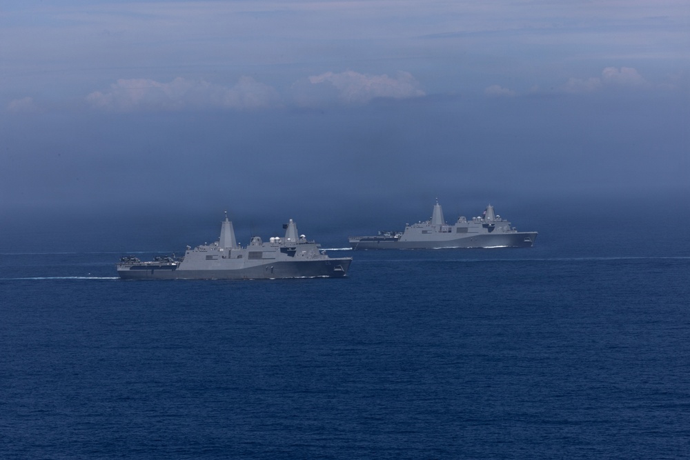 The image shows multiple gray naval warships sailing in formation across open blue ocean waters. The vessels appear to be amphibious transport dock ships (LPD-class) based on their distinctive superstructure design. The scene is captured under overcast, hazy sky conditions with low-lying clouds on the horizon.