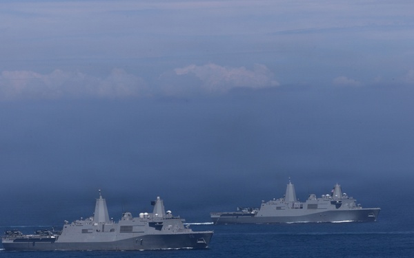 The image shows multiple gray naval warships sailing in formation across open blue ocean waters. The vessels appear to be amphibious transport dock ships (LPD-class) based on their distinctive superstructure design. The scene is captured under overcast, hazy sky conditions with low-lying clouds on the horizon.