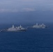 The image shows multiple gray naval warships sailing in formation across open blue ocean waters. The vessels appear to be amphibious transport dock ships (LPD-class) based on their distinctive superstructure design. The scene is captured under overcast, hazy sky conditions with low-lying clouds on the horizon.