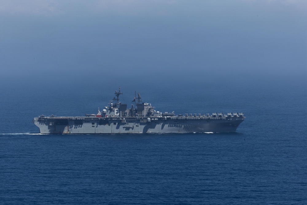 A large US Navy amphibious assault ship is underway on open ocean under overcast skies. The vessel appears to be a Wasp-class or America-class amphibious assault ship, identifiable by its flat flight deck, island superstructure, and gray hull. The ship is sailing at moderate speed with a visible bow wake.