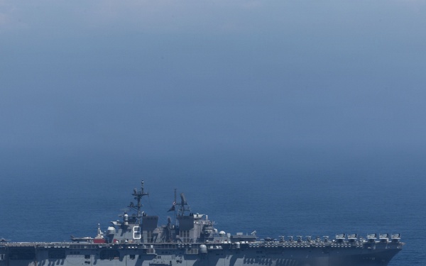 A large US Navy amphibious assault ship is underway on open ocean under overcast skies. The vessel appears to be a Wasp-class or America-class amphibious assault ship, identifiable by its flat flight deck, island superstructure, and gray hull. The ship is sailing at moderate speed with a visible bow wake.