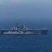 A large US Navy amphibious assault ship is underway on open ocean under overcast skies. The vessel appears to be a Wasp-class or America-class amphibious assault ship, identifiable by its flat flight deck, island superstructure, and gray hull. The ship is sailing at moderate speed with a visible bow wake.