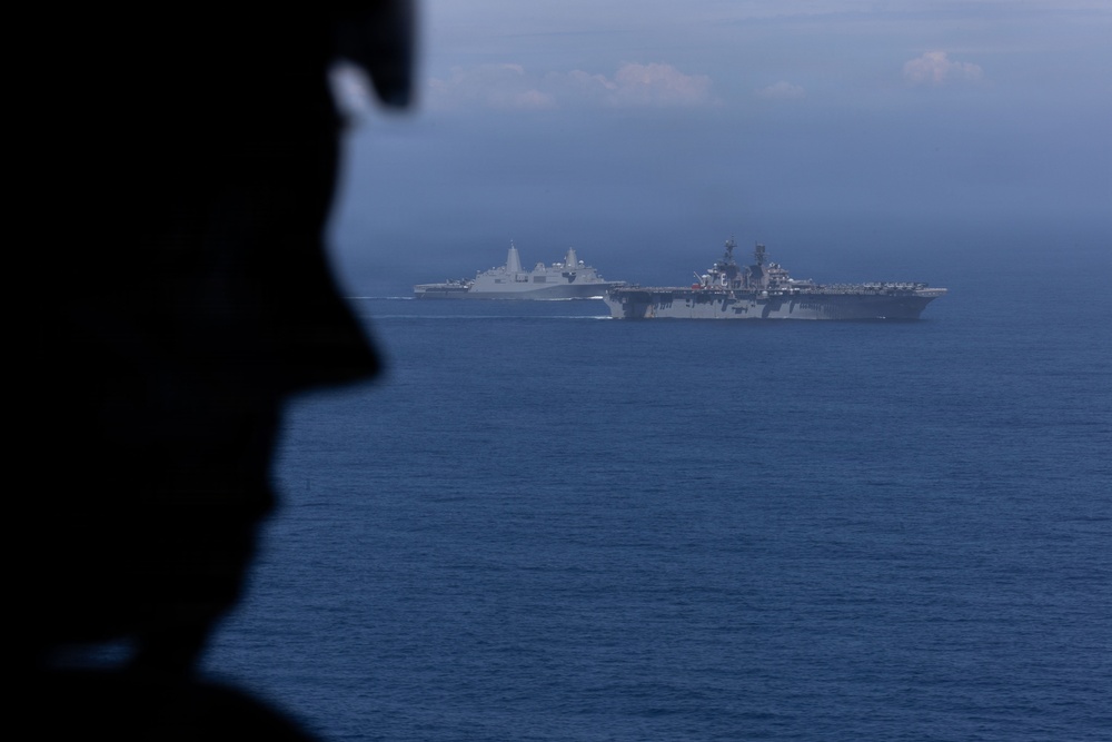 A silhouetted military personnel member is visible in the foreground, observing two large naval vessels sailing in open blue ocean waters. The scene appears to be photographed from an elevated vantage point, likely an aircraft or another vessel. The image captures what appears to be a naval formation or exercise with at least one amphibious assault ship and one amphibious transport dock vessel.