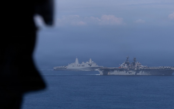 A silhouetted military personnel member is visible in the foreground, observing two large naval vessels sailing in open blue ocean waters. The scene appears to be photographed from an elevated vantage point, likely an aircraft or another vessel. The image captures what appears to be a naval formation or exercise with at least one amphibious assault ship and one amphibious transport dock vessel.