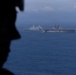A silhouetted military personnel member is visible in the foreground, observing two large naval vessels sailing in open blue ocean waters. The scene appears to be photographed from an elevated vantage point, likely an aircraft or another vessel. The image captures what appears to be a naval formation or exercise with at least one amphibious assault ship and one amphibious transport dock vessel.