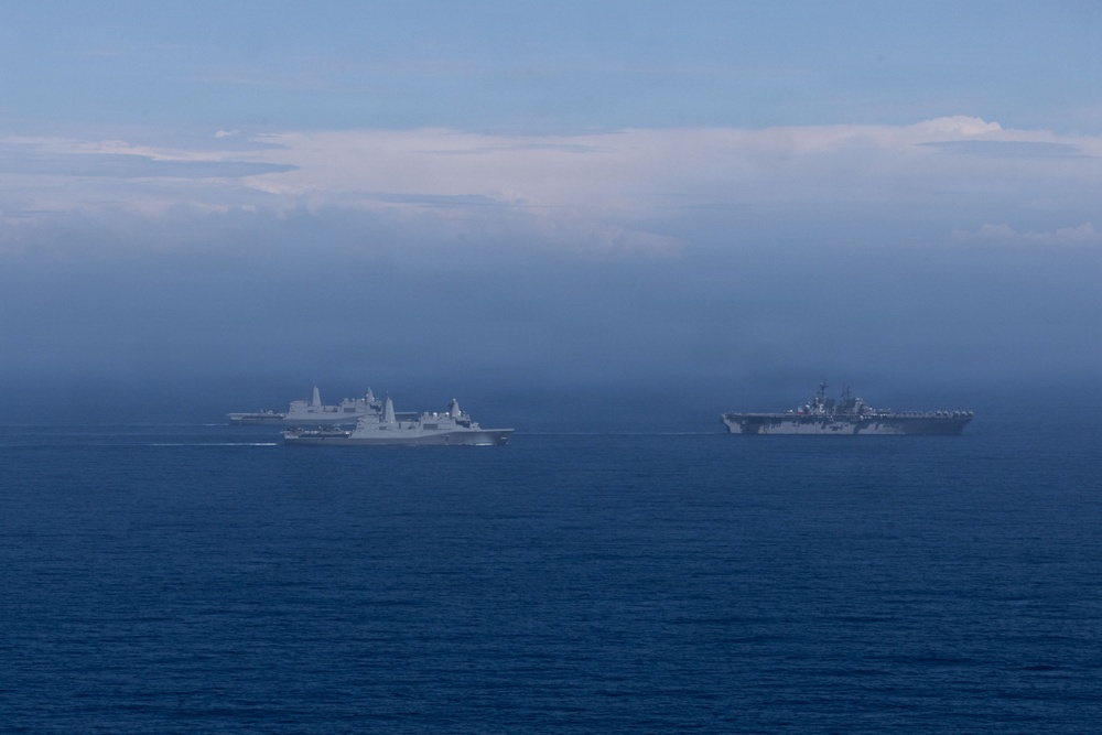 The image shows a formation of multiple naval warships underway at sea, photographed from a distance under hazy atmospheric conditions. At least three to four gray military vessels are visible, including what appears to be an amphibious assault ship or aircraft carrier on the right and transport/dock landing ships on the left. The ships appear to be conducting a coordinated naval exercise or transit operation.