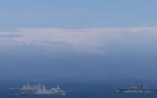 The image shows a formation of multiple naval warships underway at sea, photographed from a distance under hazy atmospheric conditions. At least three to four gray military vessels are visible, including what appears to be an amphibious assault ship or aircraft carrier on the right and transport/dock landing ships on the left. The ships appear to be conducting a coordinated naval exercise or transit operation.