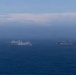 The image shows a formation of multiple naval warships underway at sea, photographed from a distance under hazy atmospheric conditions. At least three to four gray military vessels are visible, including what appears to be an amphibious assault ship or aircraft carrier on the right and transport/dock landing ships on the left. The ships appear to be conducting a coordinated naval exercise or transit operation.