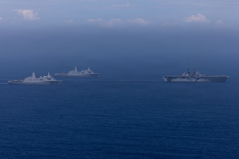 The image shows a formation of multiple naval warships sailing together on open ocean under an overcast, hazy sky. The formation appears to consist of at least four vessels, including what appears to be an amphibious assault ship (large flat-deck vessel on the right) and several San Antonio-class amphibious transport dock ships. The ships are sailing in a loose formation, suggesting a naval exercise or transit operation.