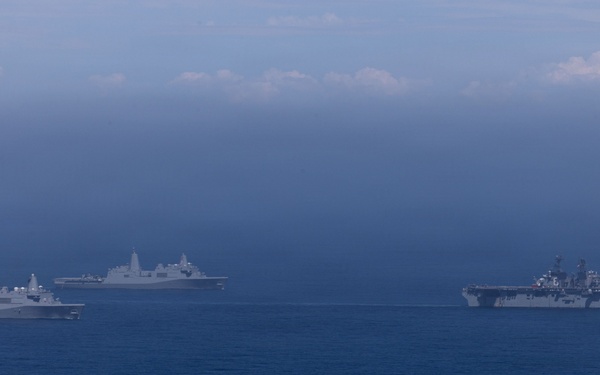 The image shows a formation of multiple naval warships sailing together on open ocean under an overcast, hazy sky. The formation appears to consist of at least four vessels, including what appears to be an amphibious assault ship (large flat-deck vessel on the right) and several San Antonio-class amphibious transport dock ships. The ships are sailing in a loose formation, suggesting a naval exercise or transit operation.