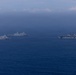 The image shows a formation of multiple naval warships sailing together on open ocean under an overcast, hazy sky. The formation appears to consist of at least four vessels, including what appears to be an amphibious assault ship (large flat-deck vessel on the right) and several San Antonio-class amphibious transport dock ships. The ships are sailing in a loose formation, suggesting a naval exercise or transit operation.