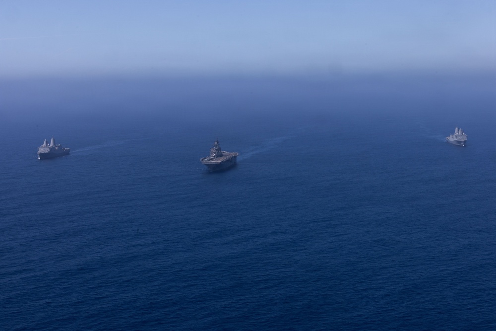 An aerial photograph shows three naval vessels sailing in formation across open deep-blue ocean water. The central vessel appears to be a large amphibious assault ship or aircraft carrier, flanked by two smaller warships on either side. The ships are leaving visible wake trails as they travel in a coordinated formation.