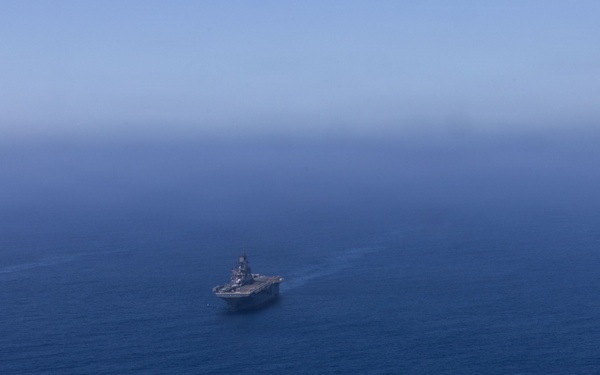 An aerial photograph shows three naval vessels sailing in formation across open deep-blue ocean water. The central vessel appears to be a large amphibious assault ship or aircraft carrier, flanked by two smaller warships on either side. The ships are leaving visible wake trails as they travel in a coordinated formation.