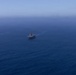 An aerial photograph shows three naval vessels sailing in formation across open deep-blue ocean water. The central vessel appears to be a large amphibious assault ship or aircraft carrier, flanked by two smaller warships on either side. The ships are leaving visible wake trails as they travel in a coordinated formation.