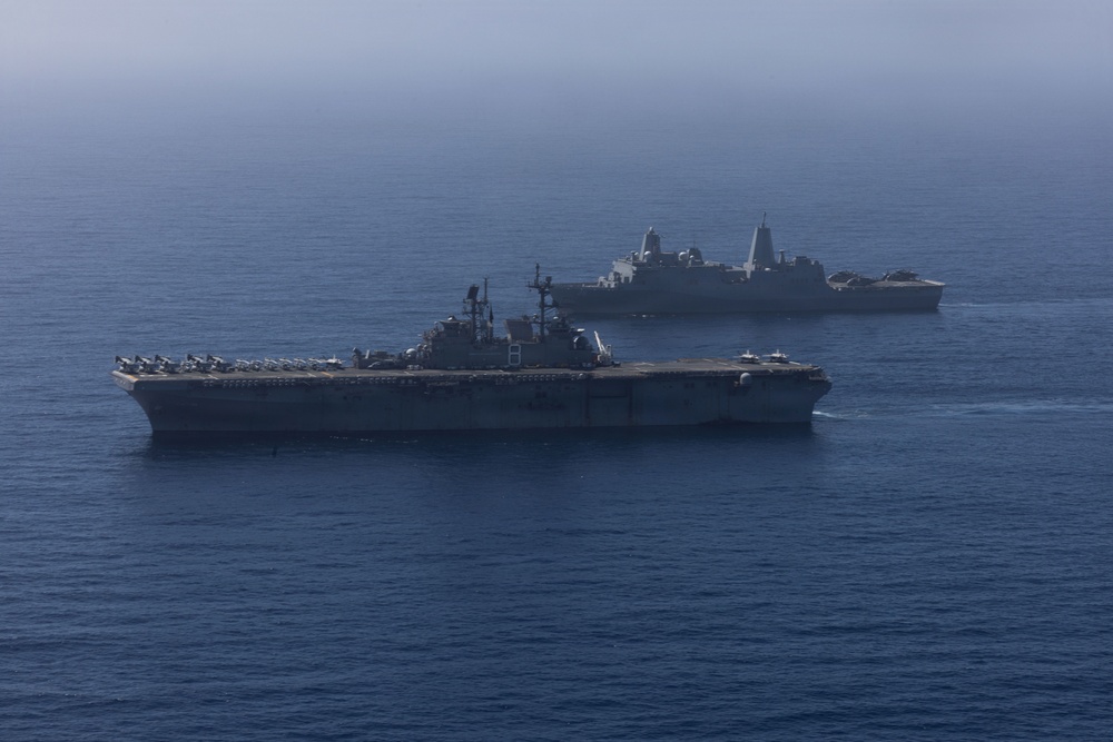 Two U.S. Navy warships are underway in open ocean under hazy atmospheric conditions. The foreground vessel is a large-deck amphibious assault ship with rotary-wing and fixed-wing aircraft visible on its flight deck. A second vessel, consistent with a San Antonio-class amphibious transport dock (LPD), is visible in the background.