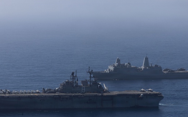 Two U.S. Navy warships are underway in open ocean under hazy atmospheric conditions. The foreground vessel is a large-deck amphibious assault ship with rotary-wing and fixed-wing aircraft visible on its flight deck. A second vessel, consistent with a San Antonio-class amphibious transport dock (LPD), is visible in the background.