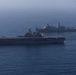 Two U.S. Navy warships are underway in open ocean under hazy atmospheric conditions. The foreground vessel is a large-deck amphibious assault ship with rotary-wing and fixed-wing aircraft visible on its flight deck. A second vessel, consistent with a San Antonio-class amphibious transport dock (LPD), is visible in the background.