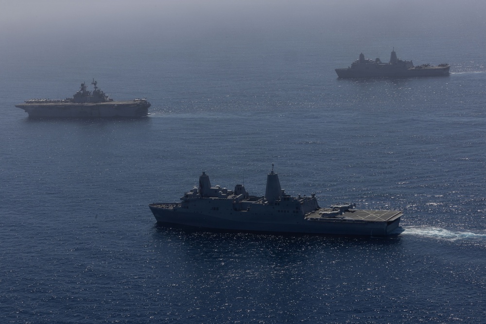 An aerial photograph shows three naval vessels underway in open blue ocean waters. The formation includes what appears to be a large amphibious assault ship or aircraft carrier in the background left, a San Antonio-class amphibious transport dock ship in the foreground center, and a third vessel in the background right. The image was taken from an elevated vantage point, likely from an aircraft, under hazy atmospheric conditions.