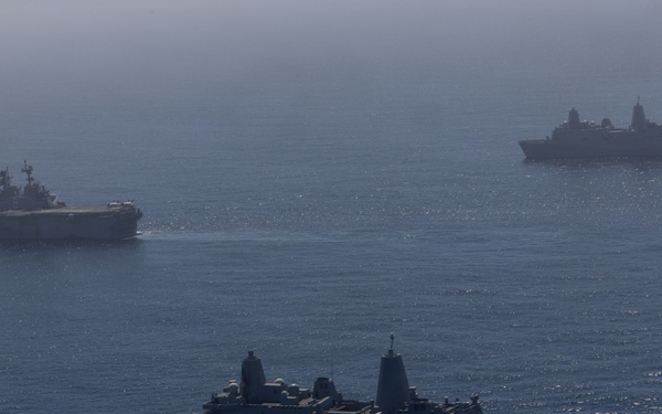 An aerial photograph shows three naval vessels underway in open blue ocean waters. The formation includes what appears to be a large amphibious assault ship or aircraft carrier in the background left, a San Antonio-class amphibious transport dock ship in the foreground center, and a third vessel in the background right. The image was taken from an elevated vantage point, likely from an aircraft, under hazy atmospheric conditions.