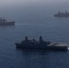 An aerial photograph shows three naval vessels underway in open blue ocean waters. The formation includes what appears to be a large amphibious assault ship or aircraft carrier in the background left, a San Antonio-class amphibious transport dock ship in the foreground center, and a third vessel in the background right. The image was taken from an elevated vantage point, likely from an aircraft, under hazy atmospheric conditions.