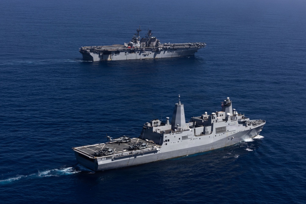 Two U.S. Navy warships are underway in open blue ocean waters. The foreground vessel appears to be a San Antonio-class amphibious transport dock (LPD) with helicopters visible on its flight deck, while the background vessel is a larger Wasp-class or America-class amphibious assault ship (LHA/LHD). Both ships are sailing in formation, suggesting a coordinated naval exercise or transit operation.
